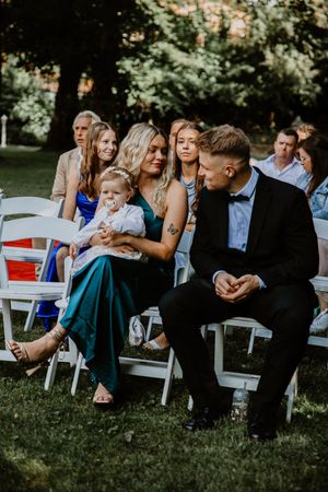 Wedding guests sit on white chairs in a garden setting during an outdoor ceremony.