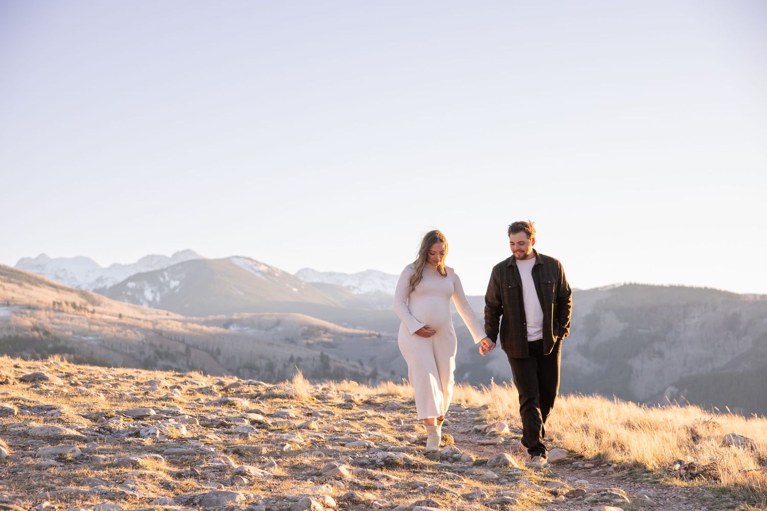 A fashionable couple in walks together along a scenic mountain ridge during golden hour for their maternity photo shoot in Vail Colorado