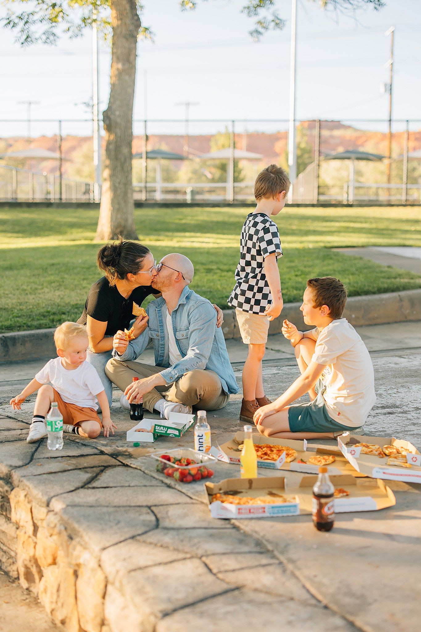 Family eating pizza while the mom and dad kiss at a park in the evening.