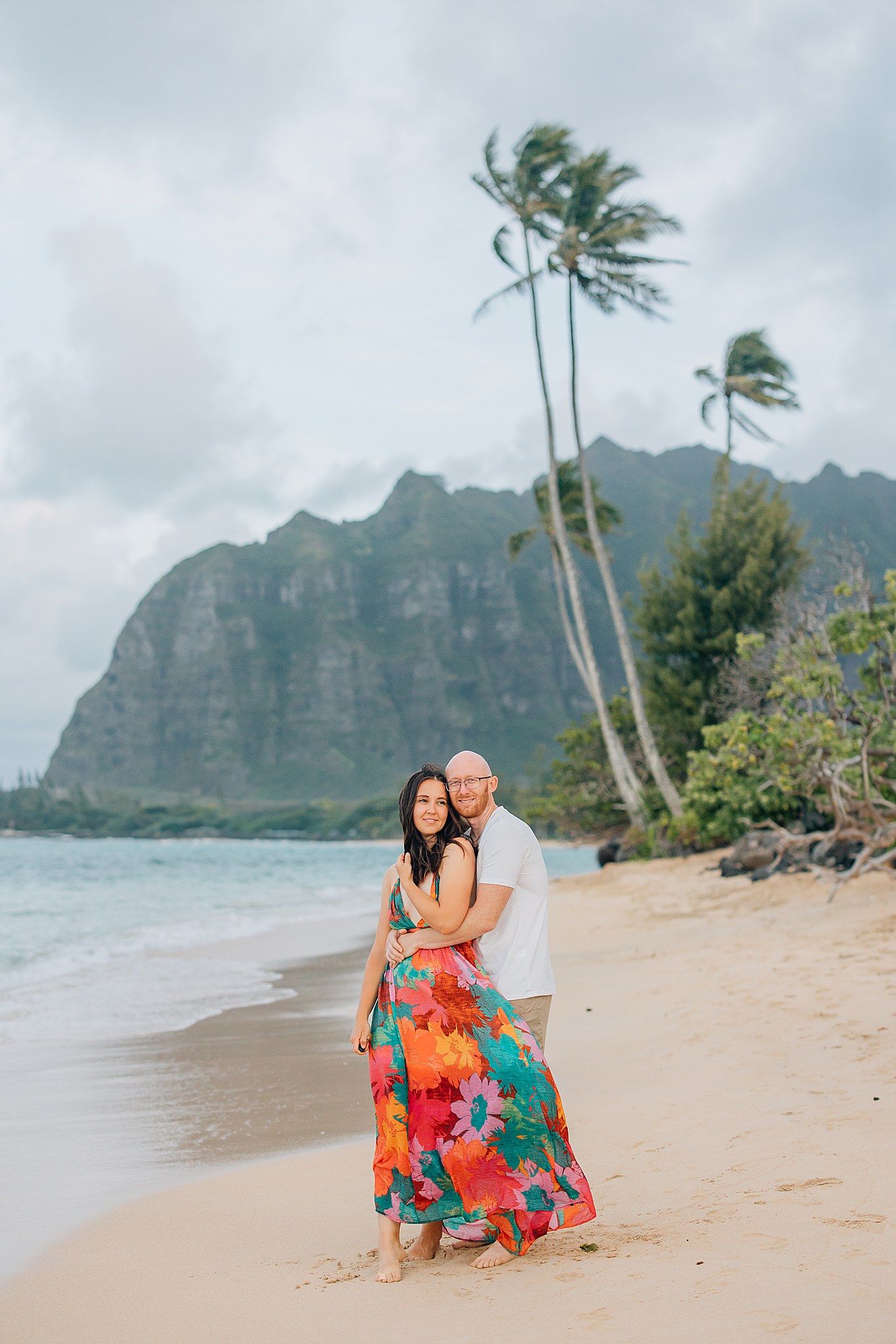 Woman and Man embrace on the beach of Oahu with mountains, ocean and palm trees in the background.