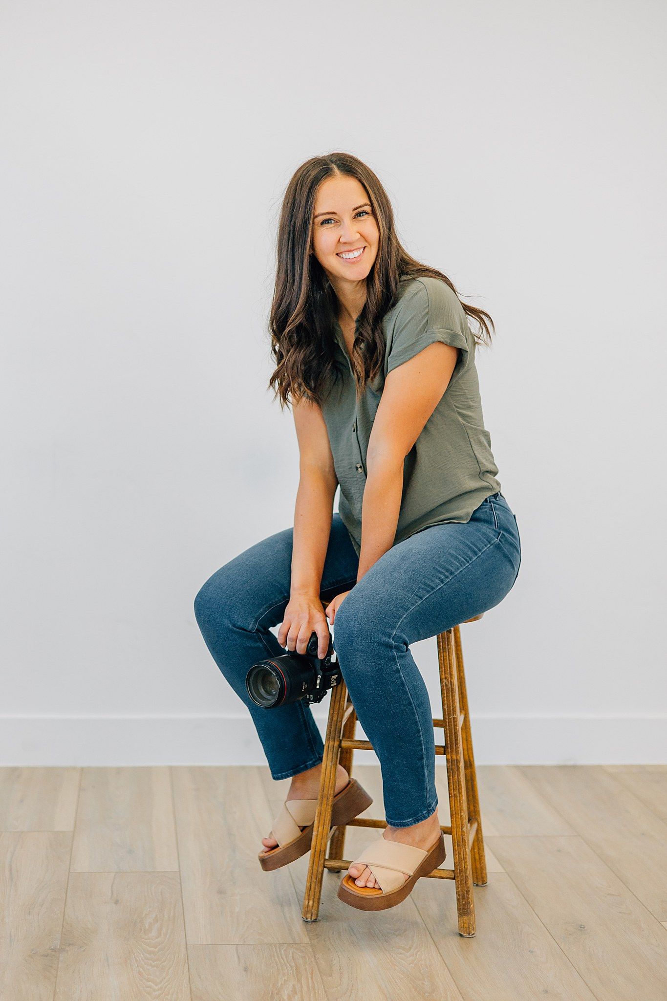 Photographer sitting on a stool, holding a camera, smiling at the camera.
