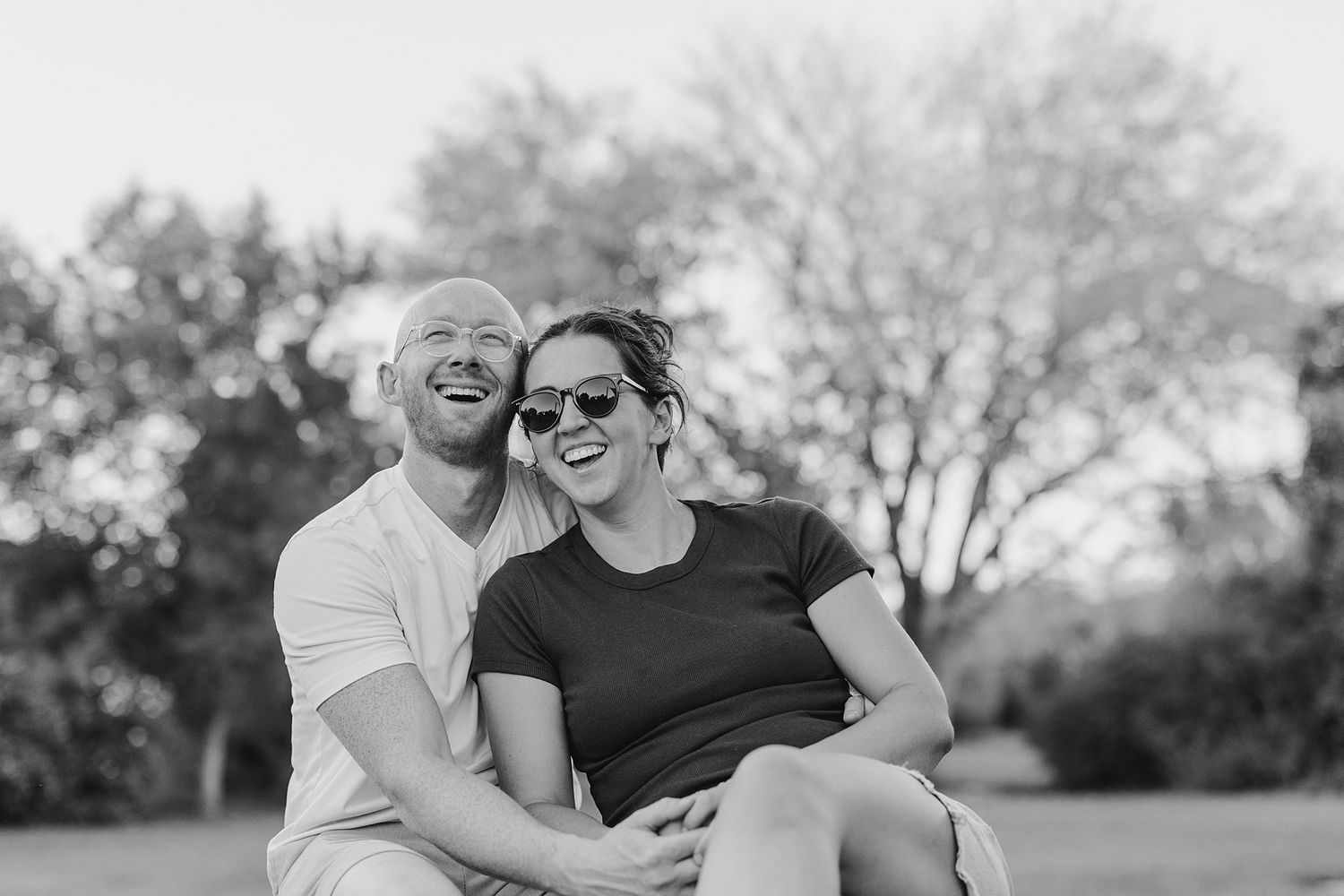 Man and woman smiling into the distance at a park. Woman has on sunglasses.