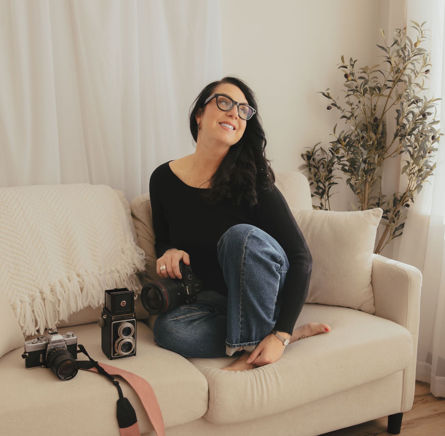 Photographer in black sweater and jeans lounges on white couch with vintage camera and decorative plant in background.