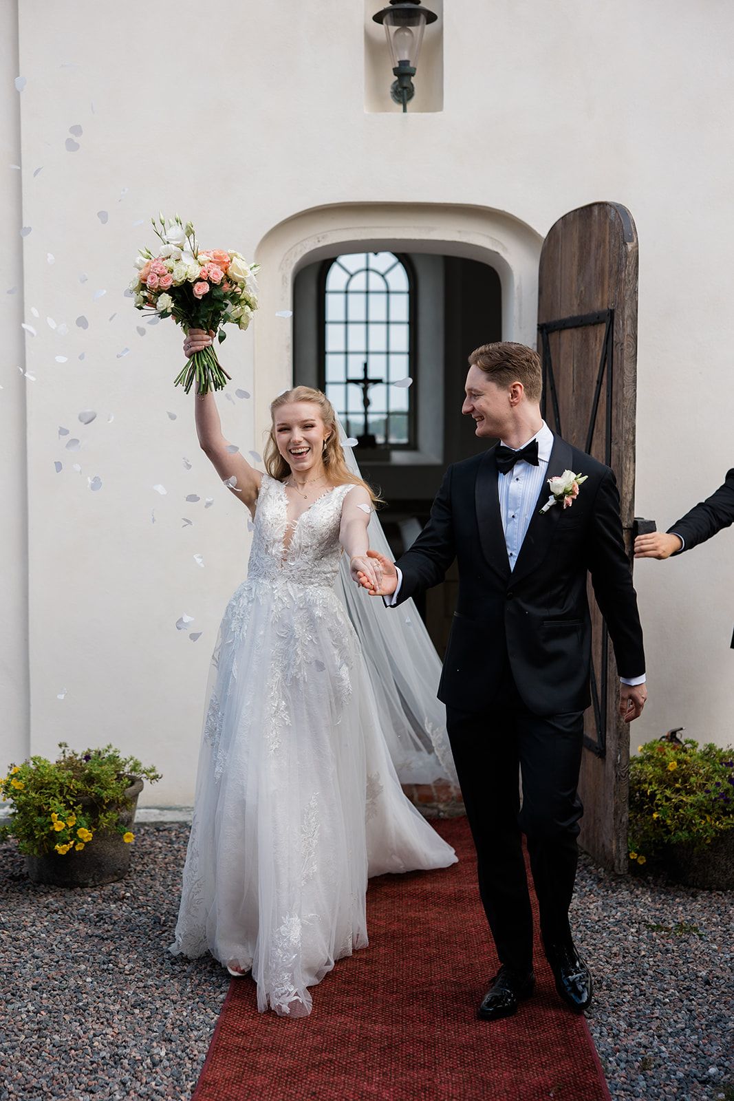 A couple in wedding attire walks together on a gravel path outside a historic stone building with arched doorways.