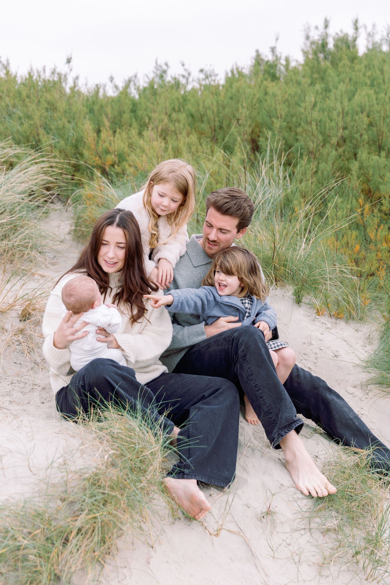 A family shares tender moments together while relaxing on sandy dunes surrounded by coastal grass on a beach.