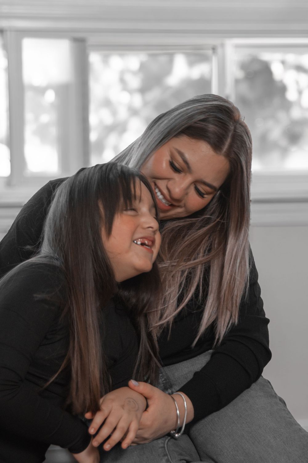 Two people in black clothing embrace and laugh together in a bright interior setting.