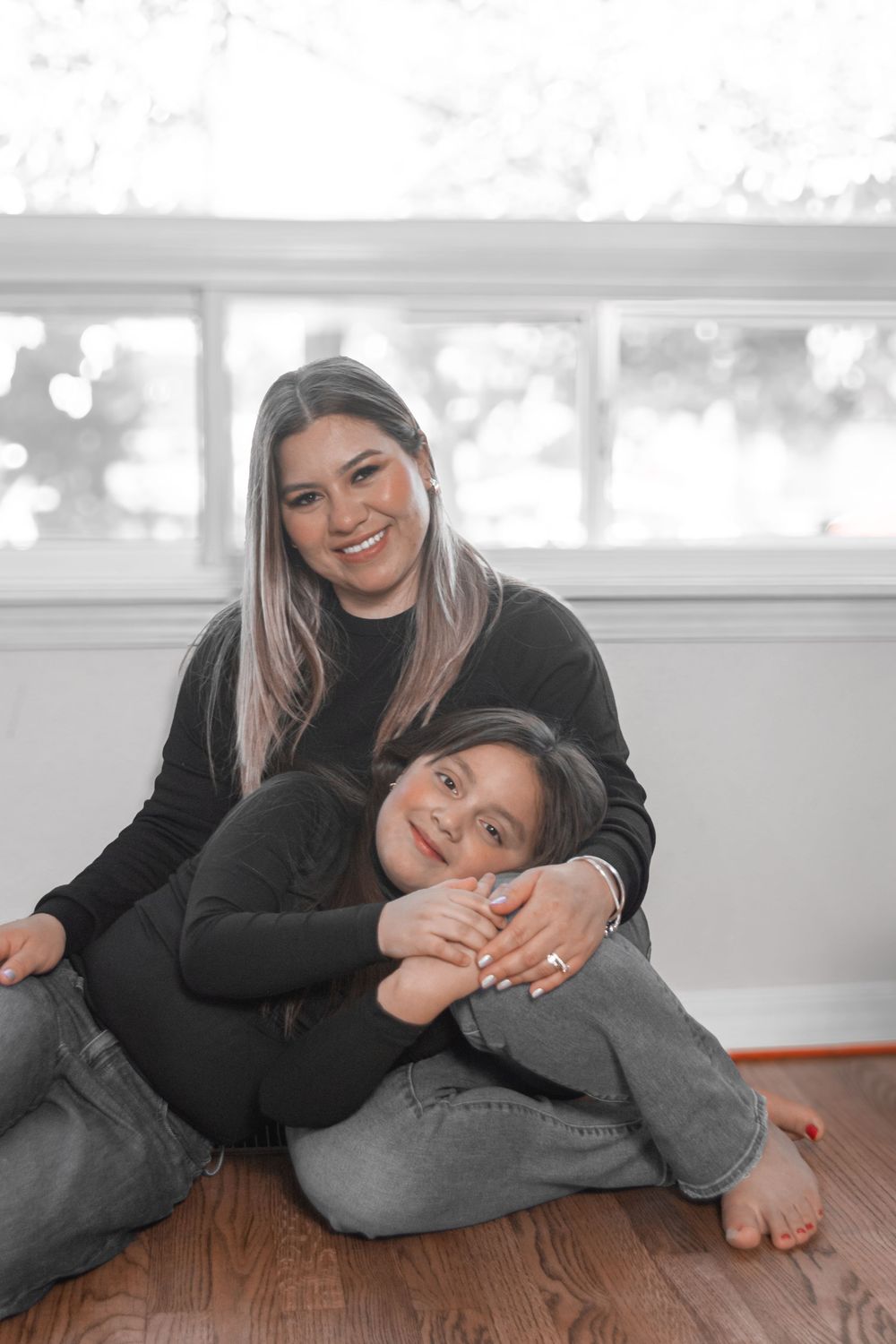 A parent and child share a tender moment sitting together on a wooden floor in matching black sweaters.