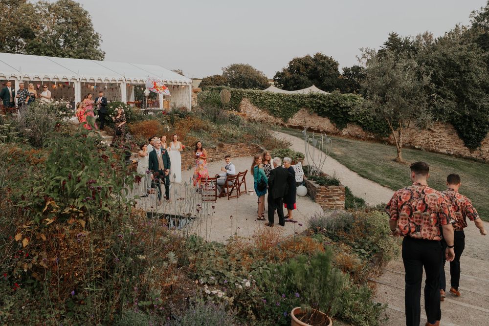 Guests socializing in a beautiful garden wedding venue with lush greenery and a white marquee tent.