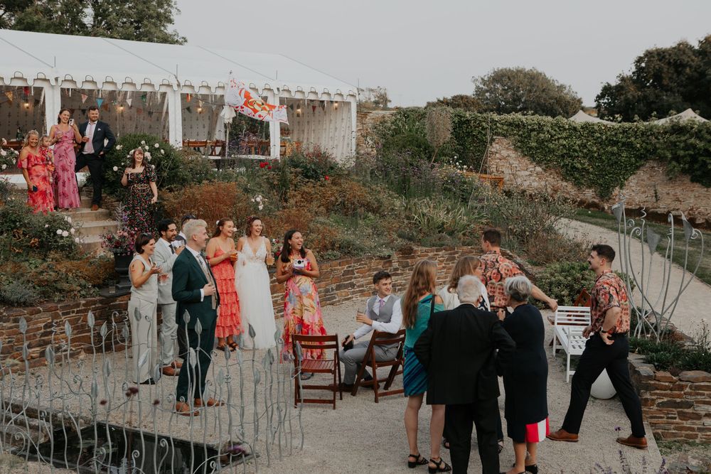 Wedding guests mingling outdoors in a terraced garden with a white marquee tent in the background.