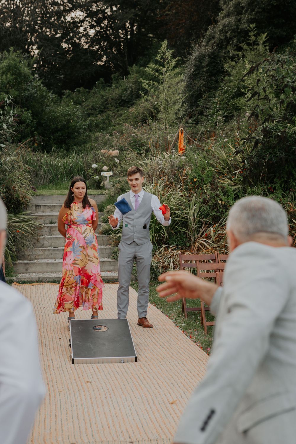 Young couple playing cornhole lawn game at an outdoor garden wedding reception with guests watching.