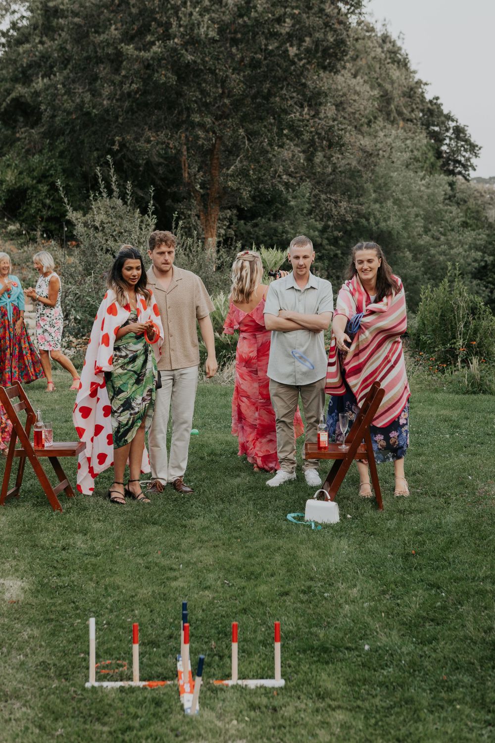 Wedding guests wrapped in colorful blankets playing lawn games on grass at an outdoor garden reception.