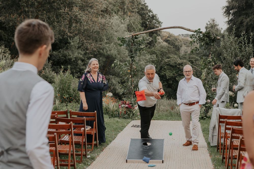 Wedding guests playing cornhole bean bag toss game on a wooden board at an outdoor garden reception.