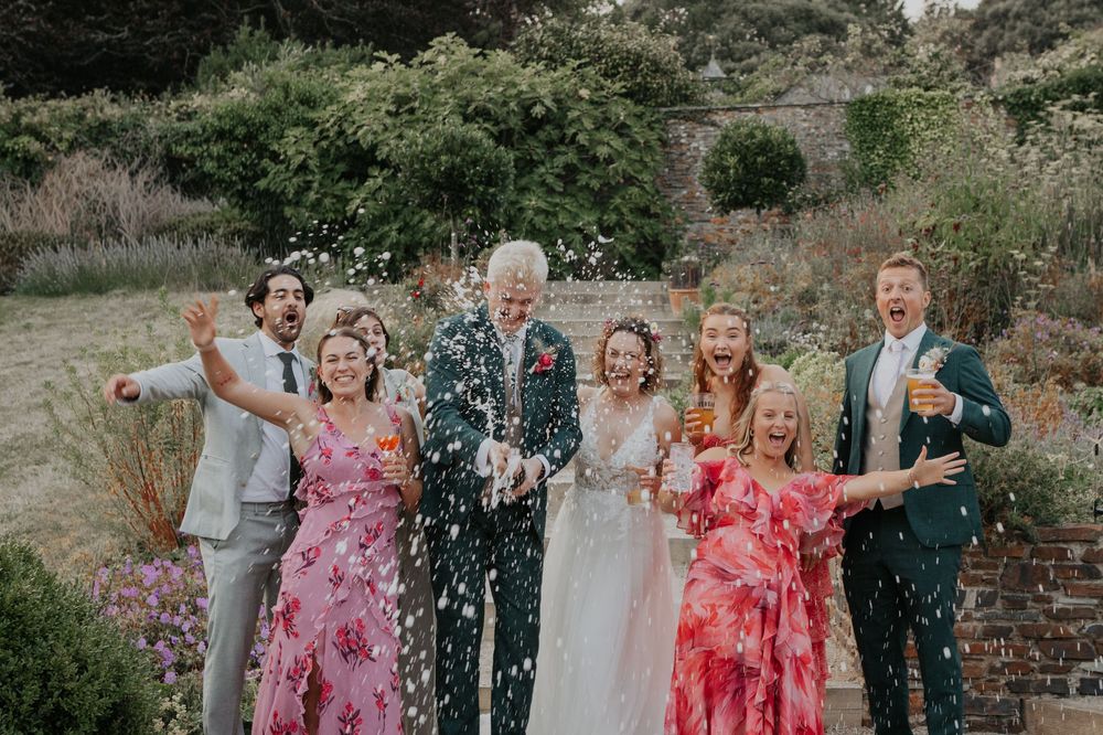 Wedding party celebrating with confetti in a garden, bride in white gown surrounded by colorful dressed guests.