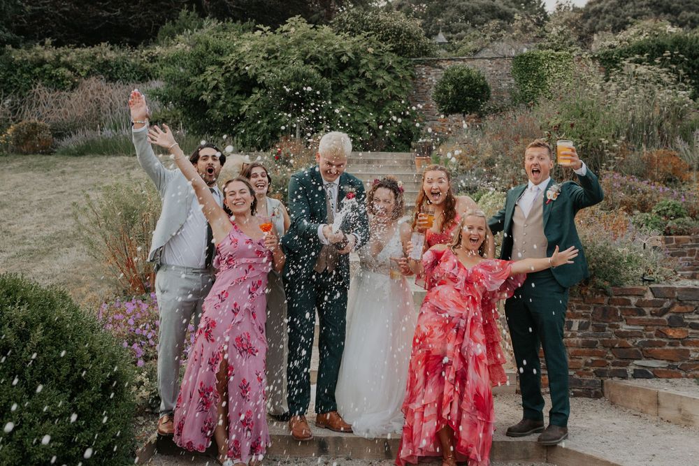 Excited wedding party celebrating with confetti shower outdoors, bride in white surrounded by guests in pink dresses.