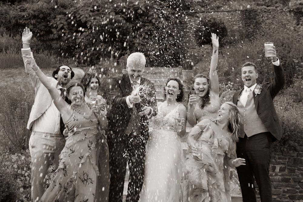 Black and white photo of joyful wedding party throwing confetti with bride and groom in a garden.