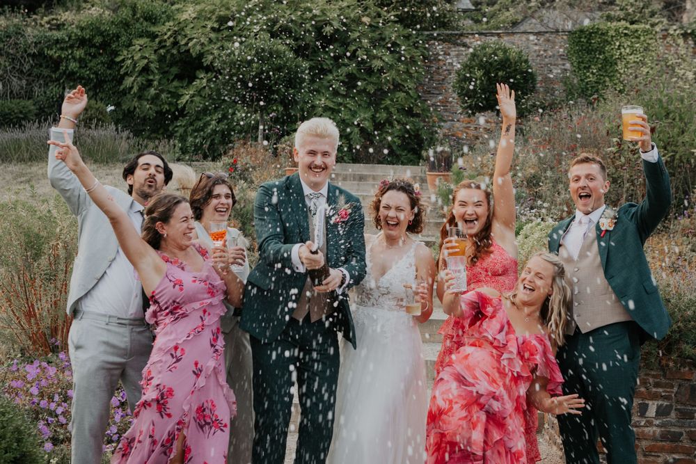 Wedding party celebrates with champagne spray in a colorful garden, bride and groom surrounded by joyful guests.