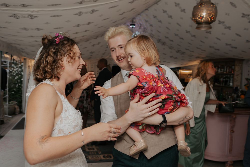 Smiling bride and groom hold a laughing baby girl at their elegant wedding reception celebration.