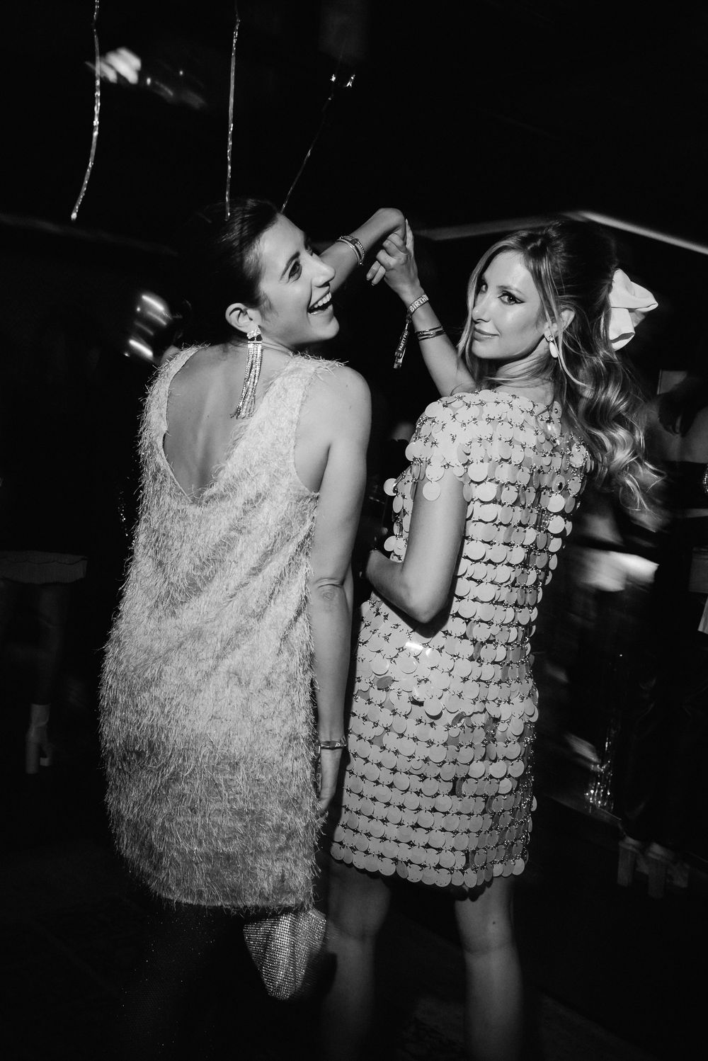 Two partygoers in shimmering dresses dance together at a nightclub in black and white photography.