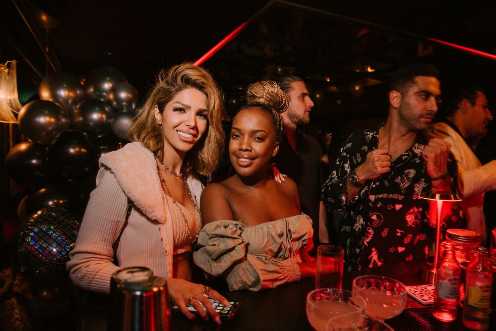 Two friends enjoying drinks and smiling together at a dimly lit nightclub with red ambient lighting.