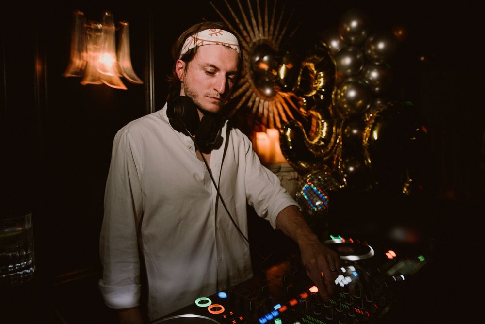 DJ in white shirt and headphones performing at nightclub with warm lighting and decorative elements in background.