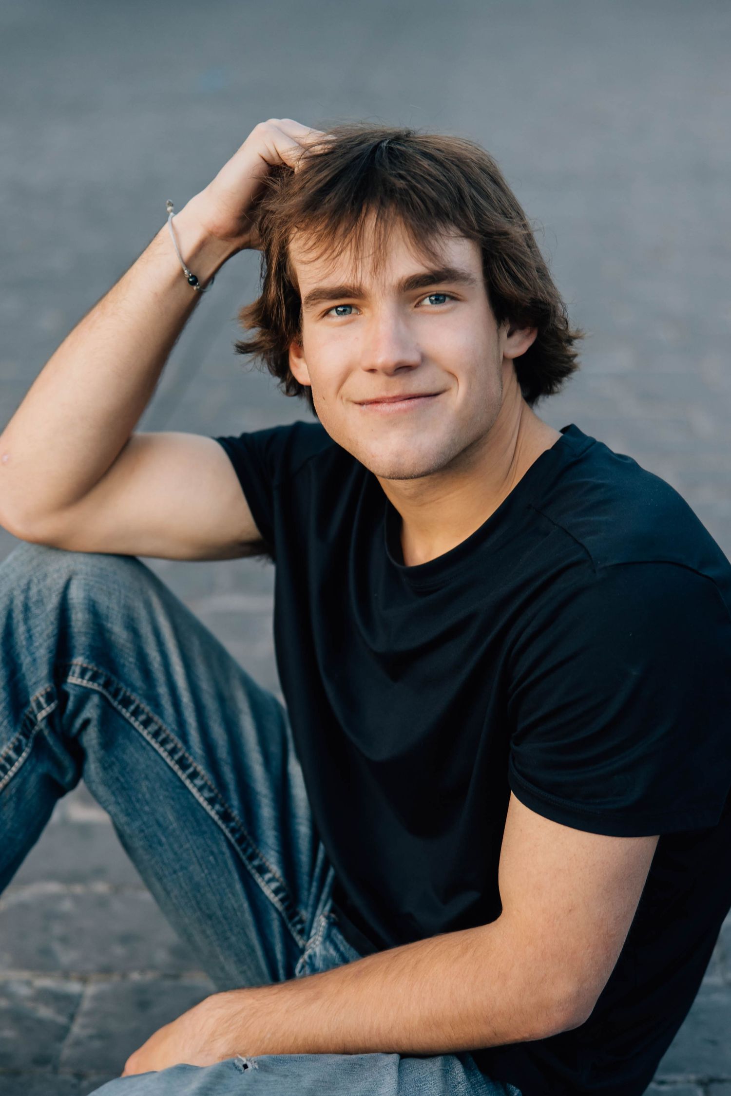 Smiling teenage boy in black t-shirt sitting outdoors with hand in hair.