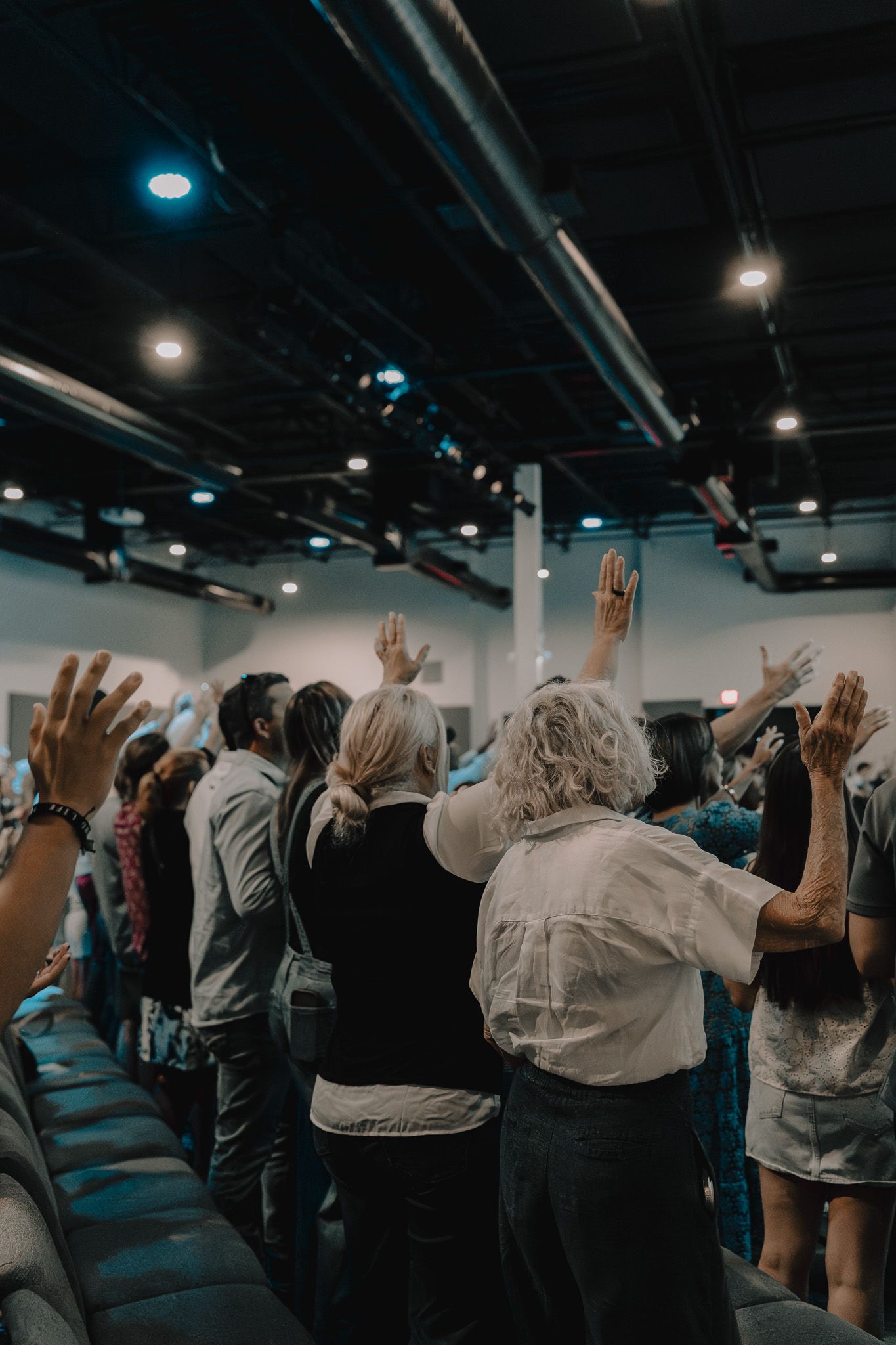 A group of people raise their hands in worship during a church service in a dimly lit auditorium.