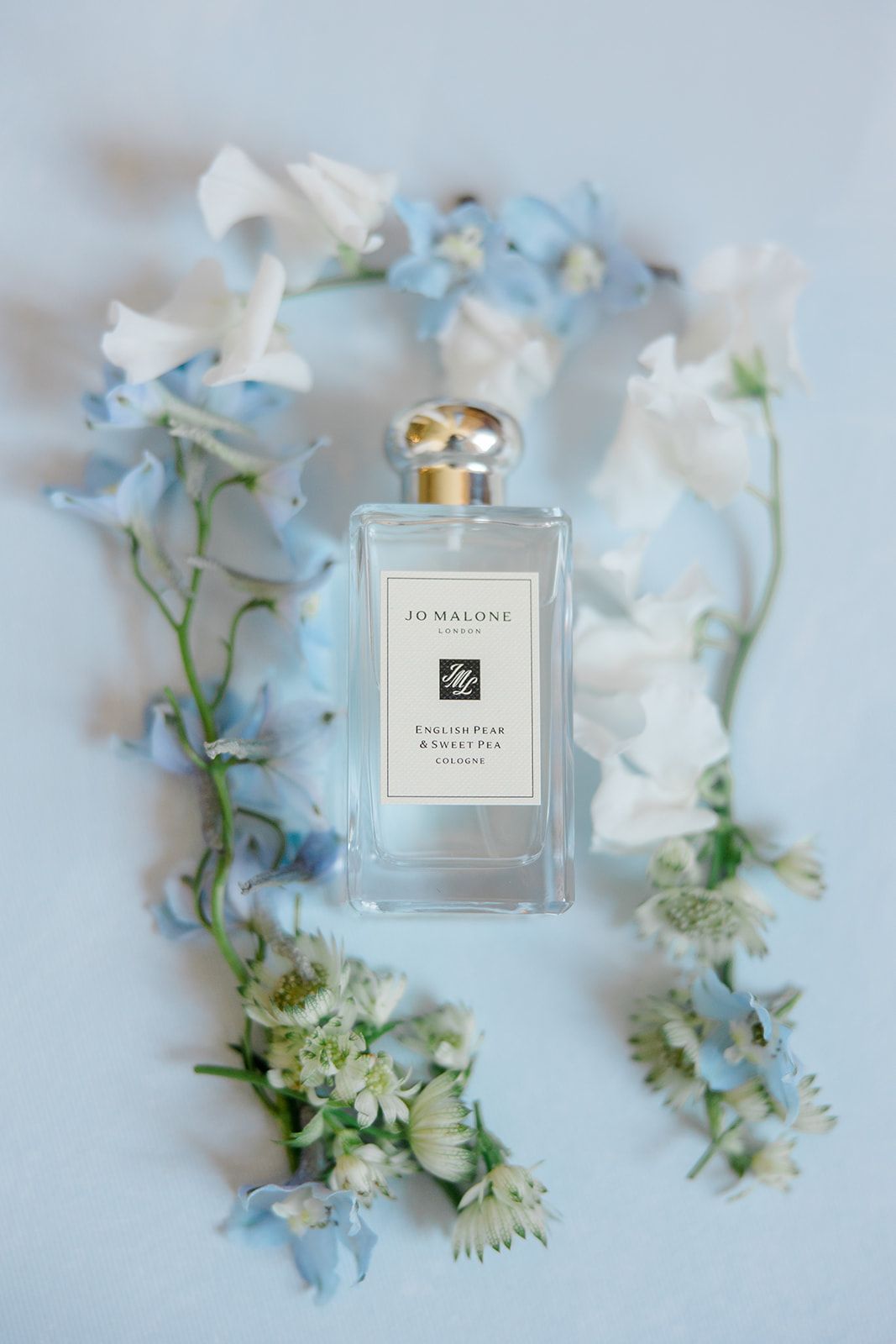 Jo Malone perfume bottle surrounded by delicate blue and white flowers on a soft gray background.