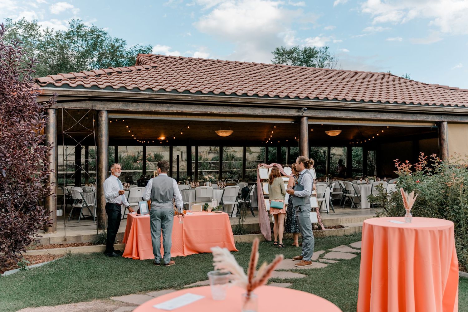 Outdoor wedding reception venue with orange tablecloths and covered pavilion surrounded by lush green landscaping.