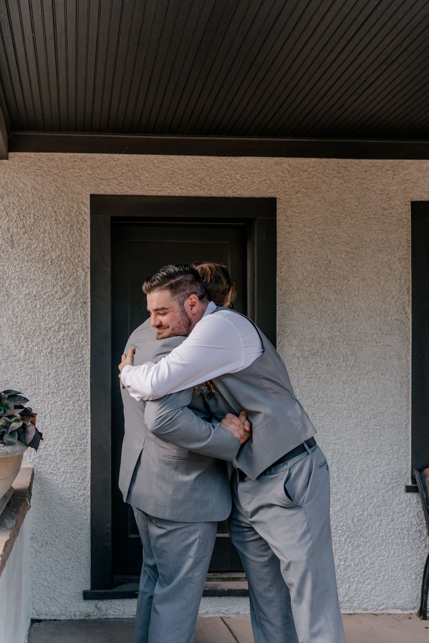 Two people in gray suits embrace in an emotional moment outside a building with white walls.