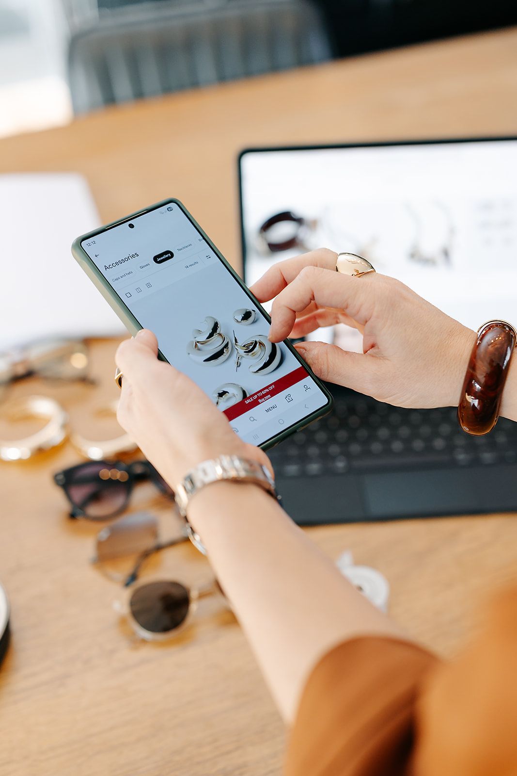 Person using smartphone while working at wooden desk with laptop and glasses in background.