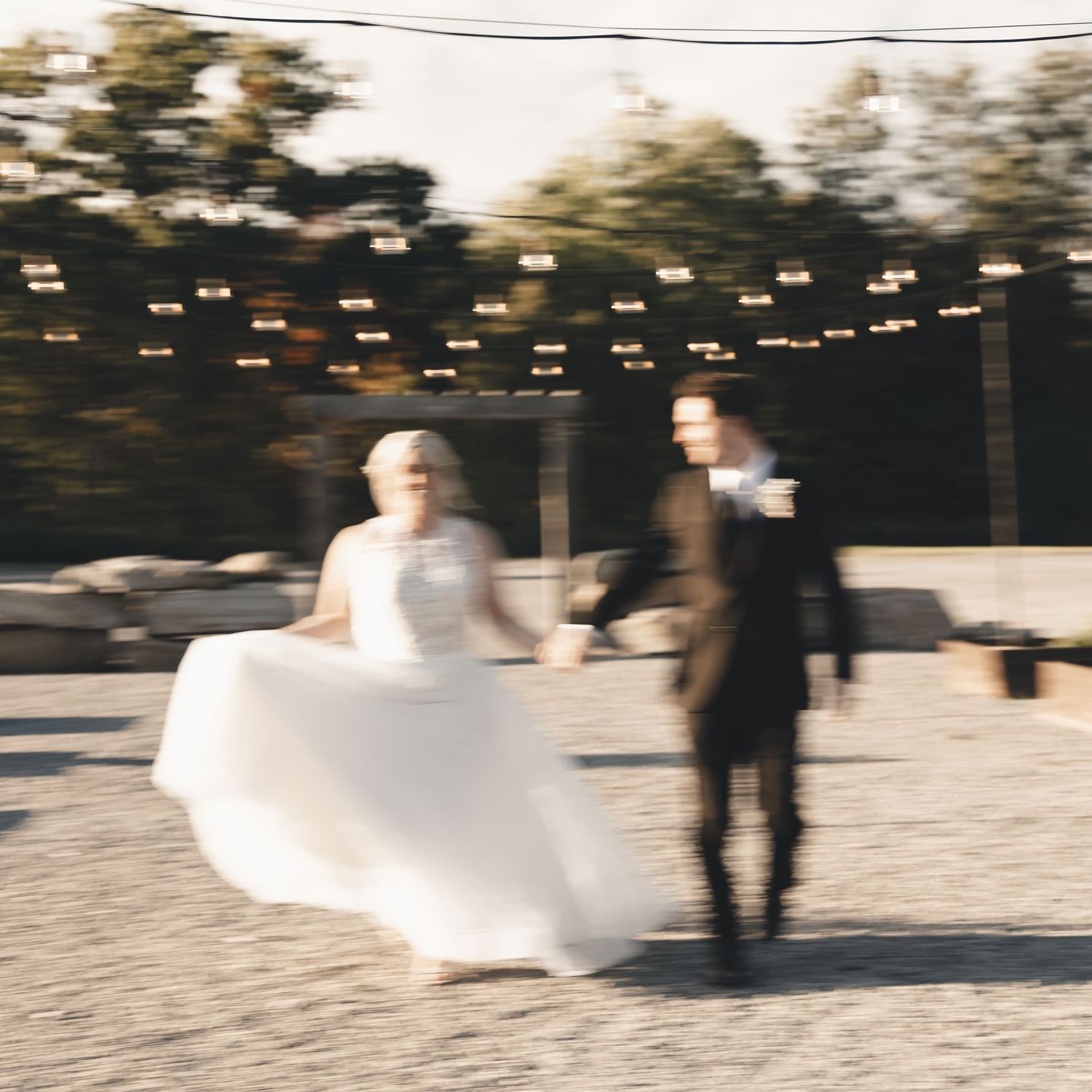 A joyful, blurred moment of a bride and groom in motion, hand in hand beneath glowing string lights on a golden evening.