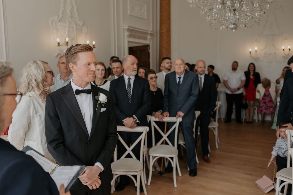 Groom in black tuxedo stands at altar watching wedding procession in elegant chandelier-lit ceremony room.
