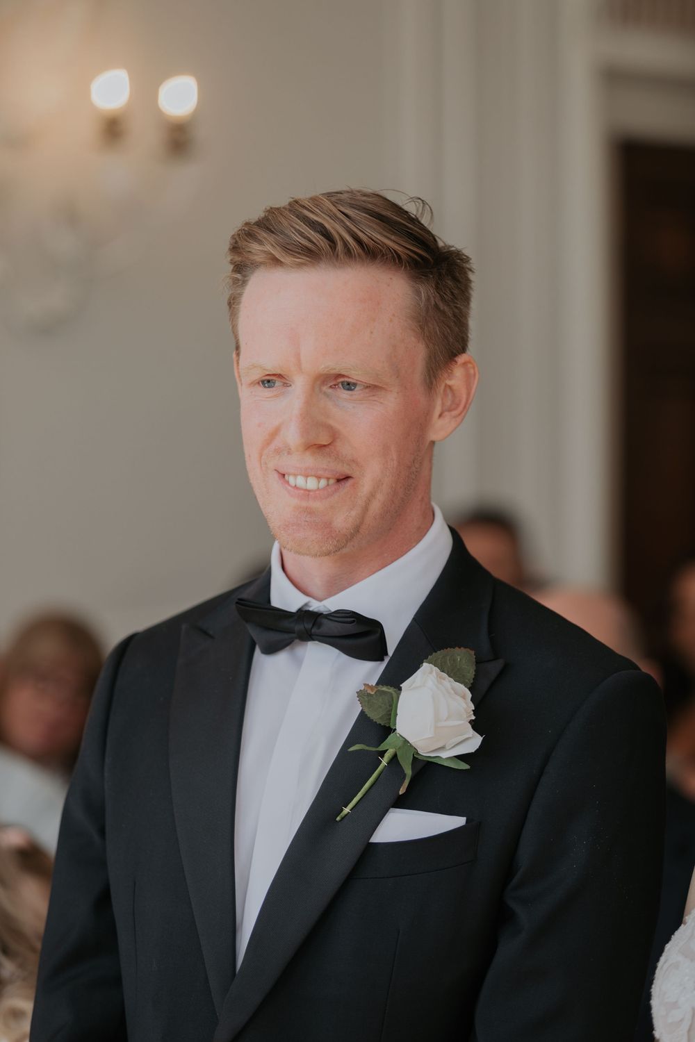 Close-up of smiling groom in black tuxedo with white rose boutonniere during wedding ceremony.