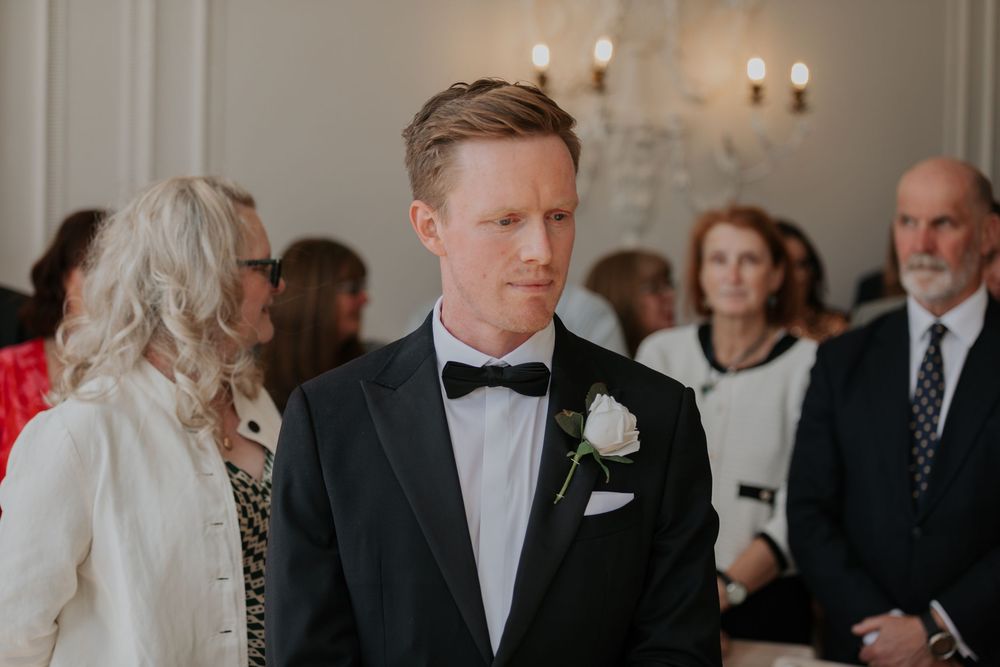Groom in black tuxedo with white rose boutonniere stands attentively during wedding ceremony.
