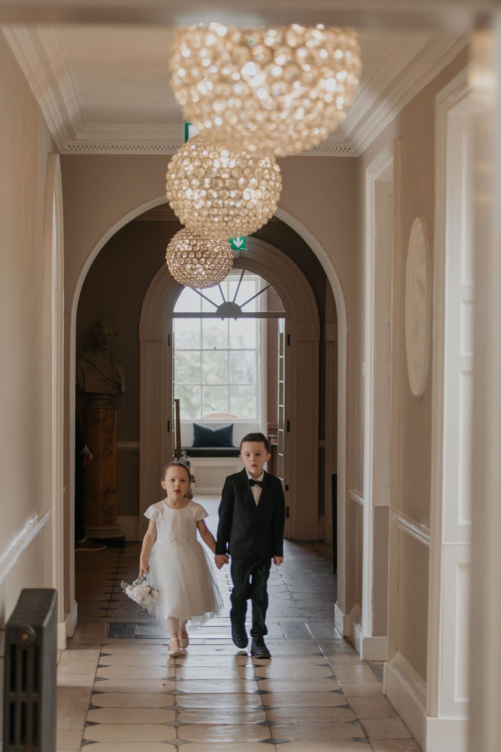 Flower girl and ring bearer in formal attire walk hand-in-hand down hallway beneath crystal chandeliers.