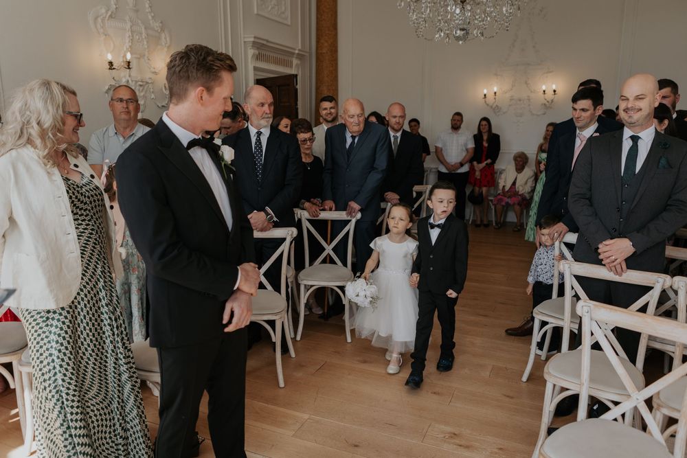 Groom in black tuxedo smiles as flower girl and ring bearer walk down wedding ceremony aisle.