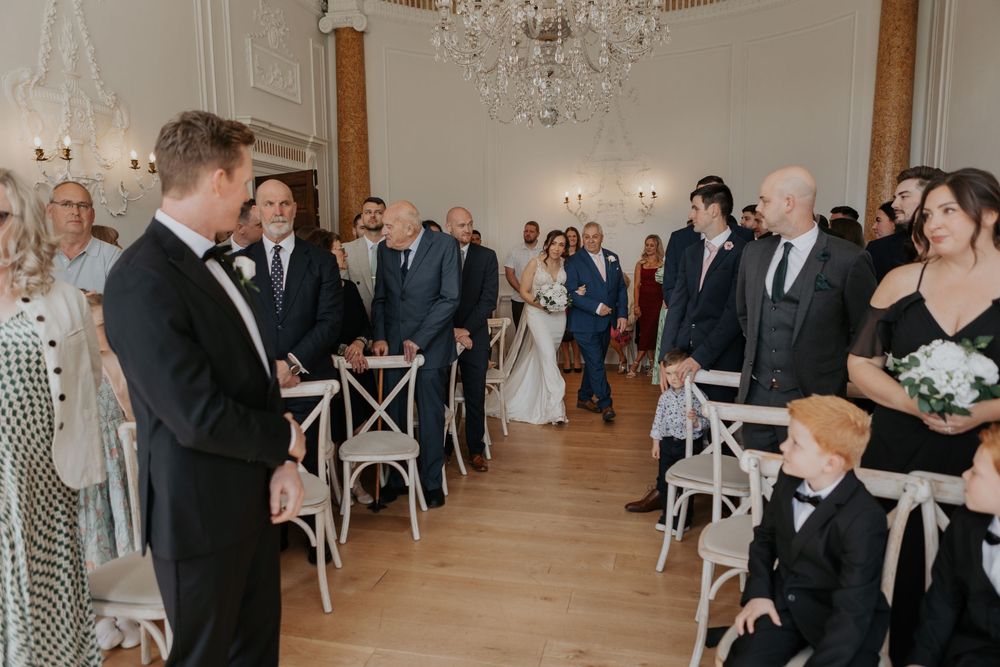 Bride walks down the aisle with escort at elegant wedding ceremony in ornate white venue.
