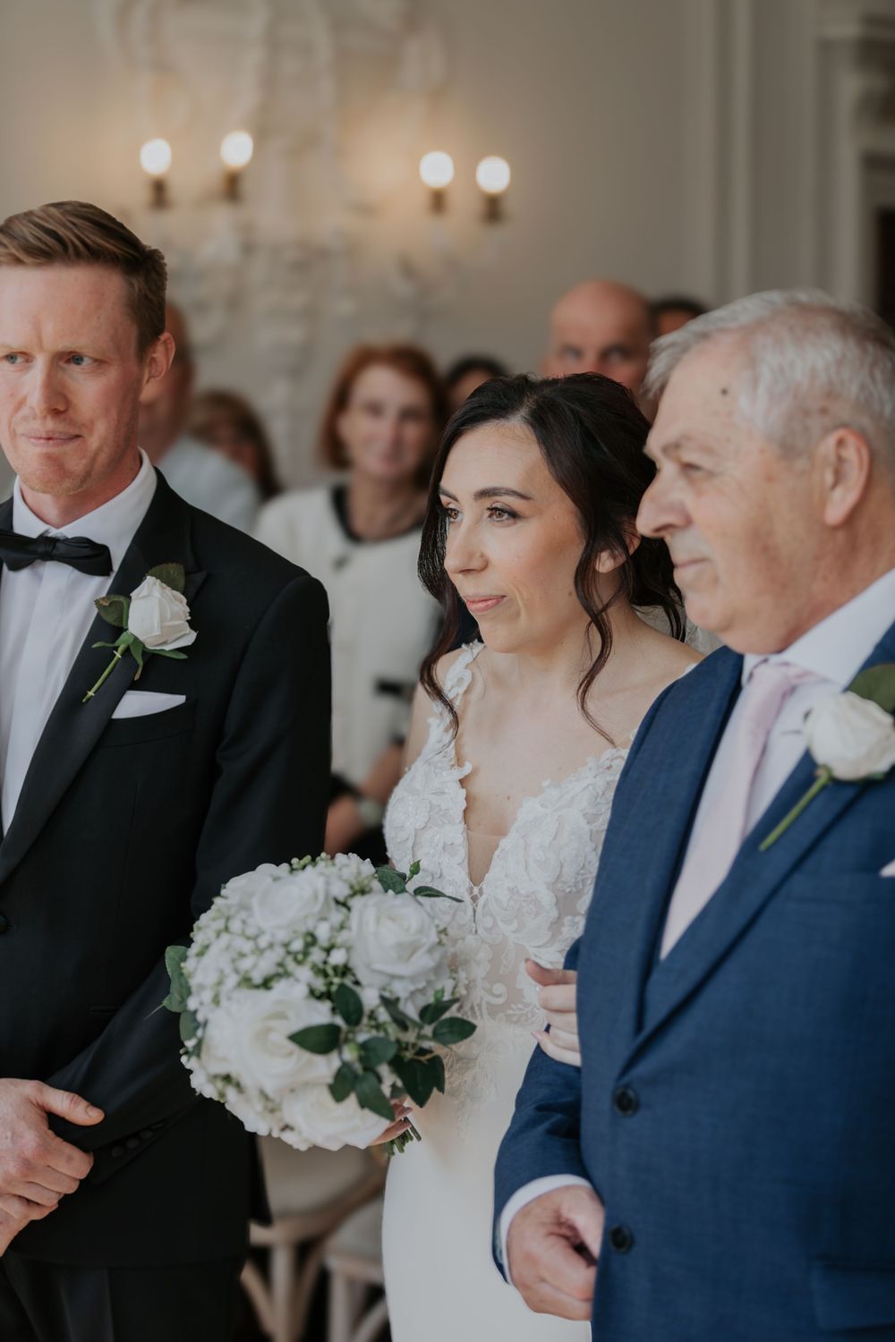 Bride in lace gown holds white bouquet while walking down aisle with father at ceremony.