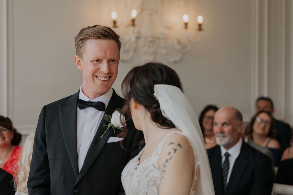 Groom in black tuxedo smiles lovingly at bride during intimate wedding ceremony exchange.