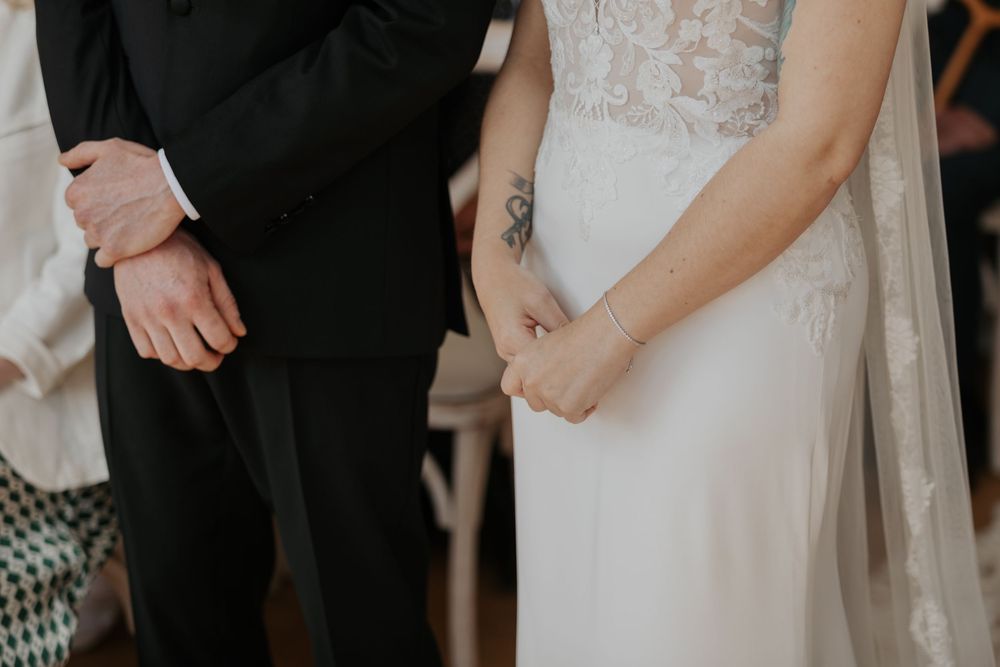 Close-up of bride and groom holding hands during wedding ceremony, bride has floral tattoo.