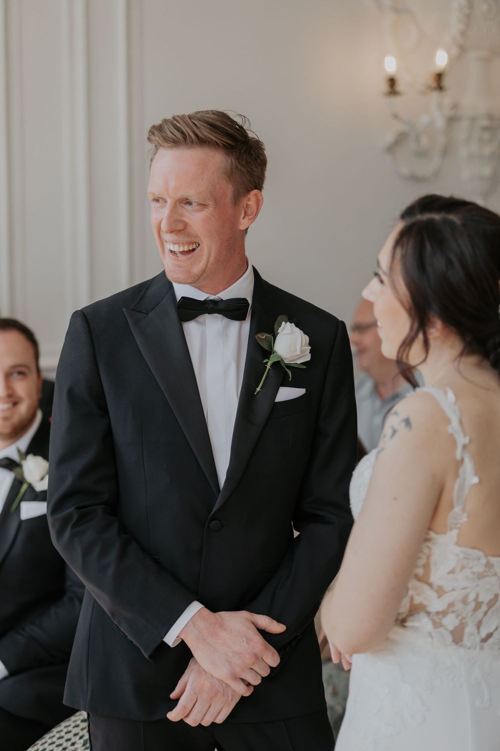 Groom in black tuxedo laughs joyfully while facing bride during elegant wedding ceremony.