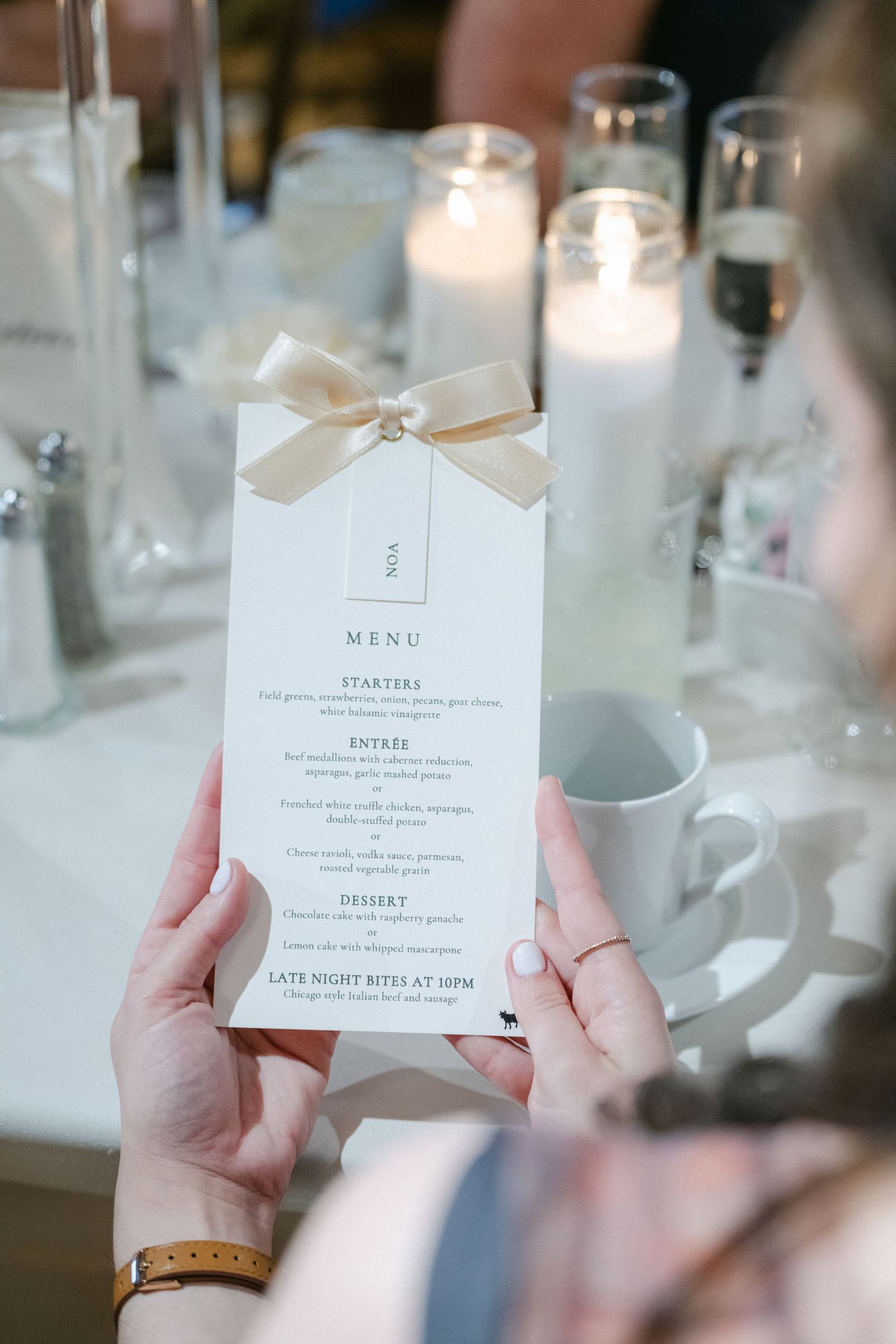 Hands holding an elegant wedding menu card with cream colored paper and delicate ribbon detail at a formal table setting.