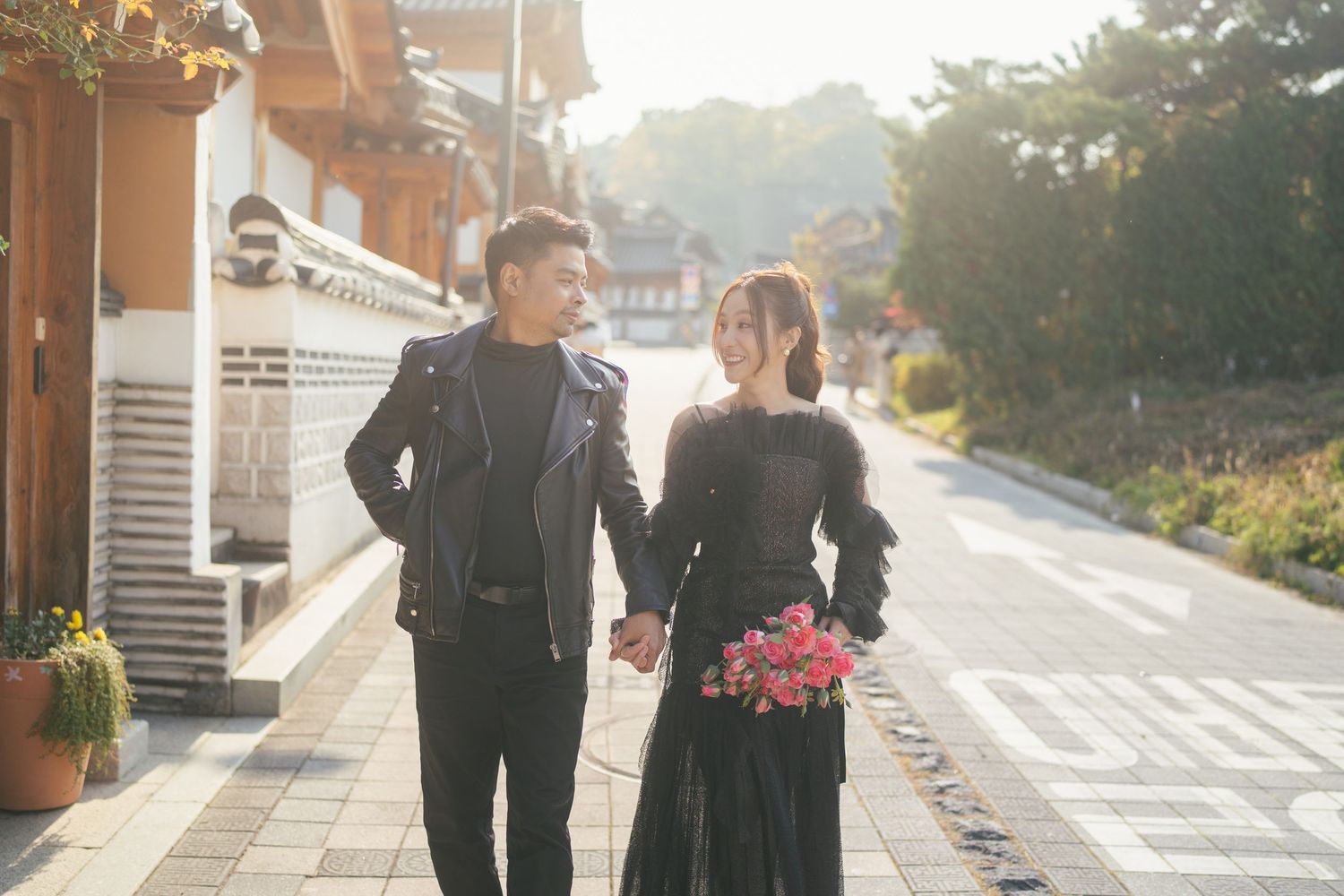 A couple in formal attire walks along a sunlit path outside a traditional Korean building at sunset.