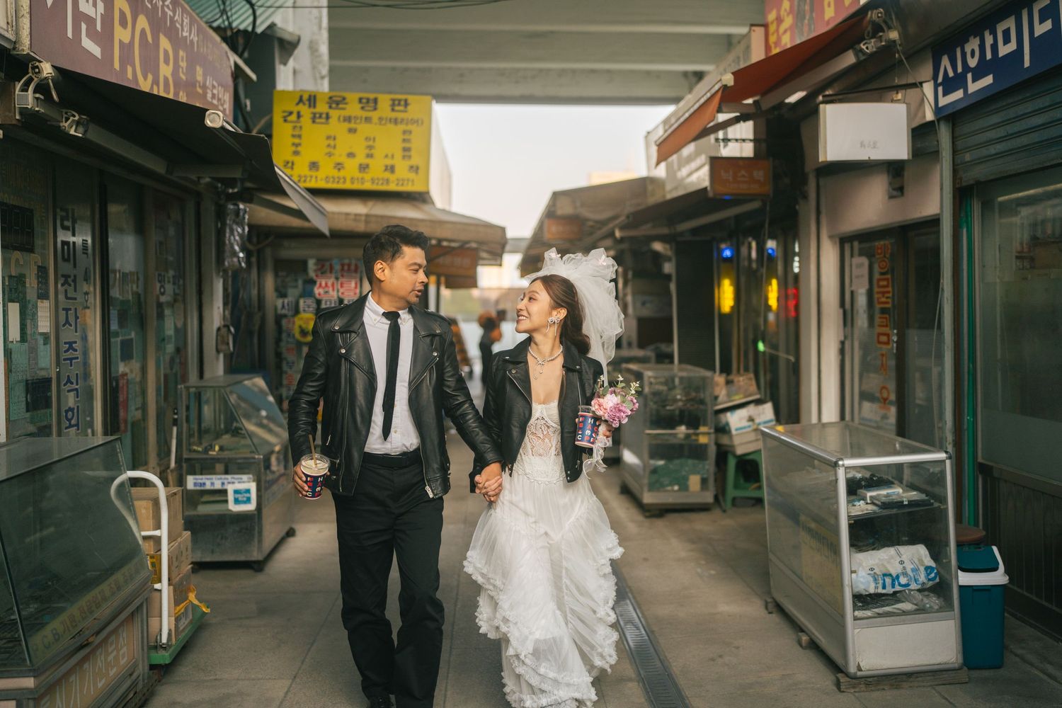 A wedding couple walks together through a narrow alley lined with traditional Korean storefronts and shops.