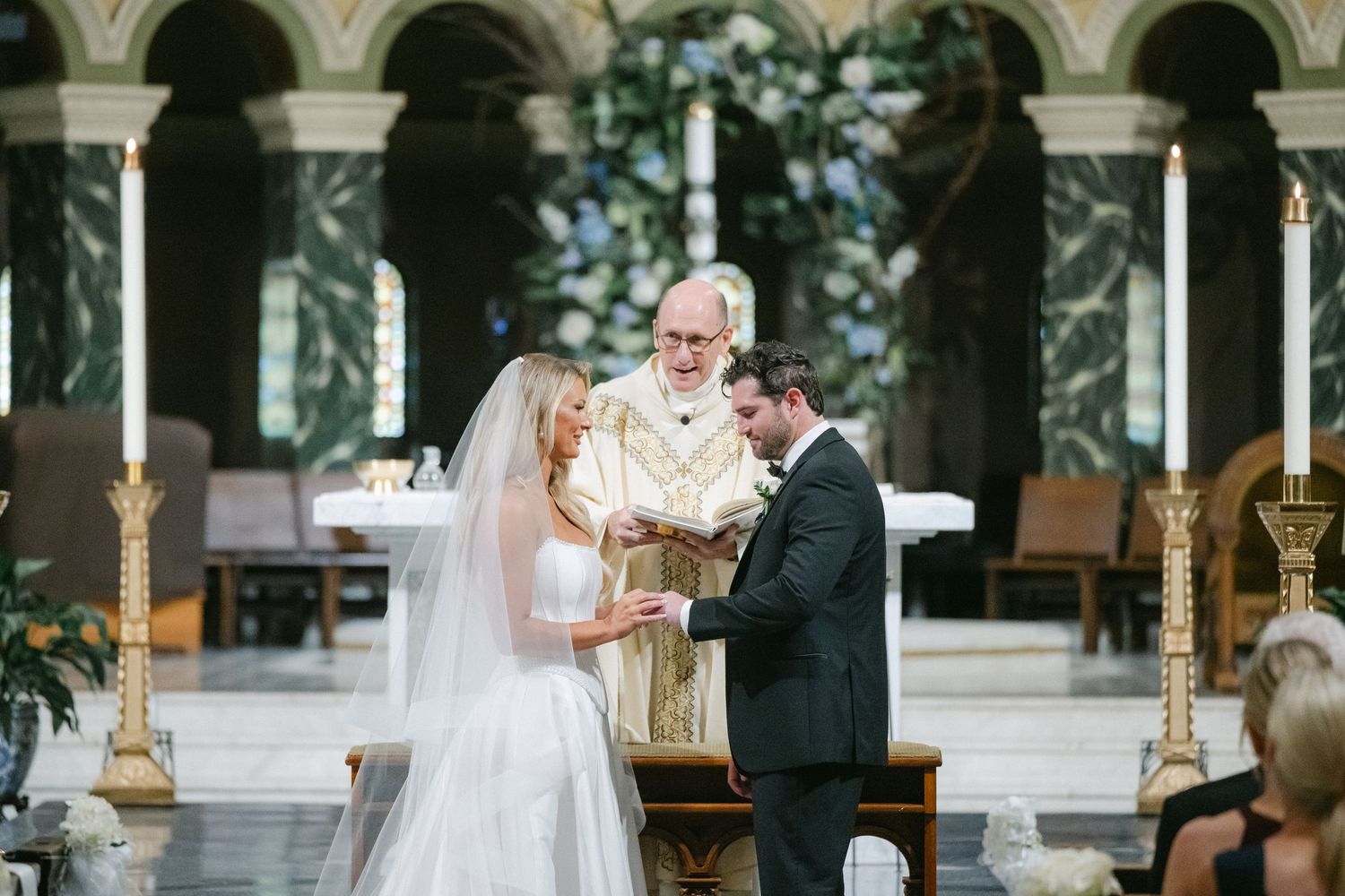 A wedding ceremony takes place in an elegant venue with white floral arrangements and decorative columns.