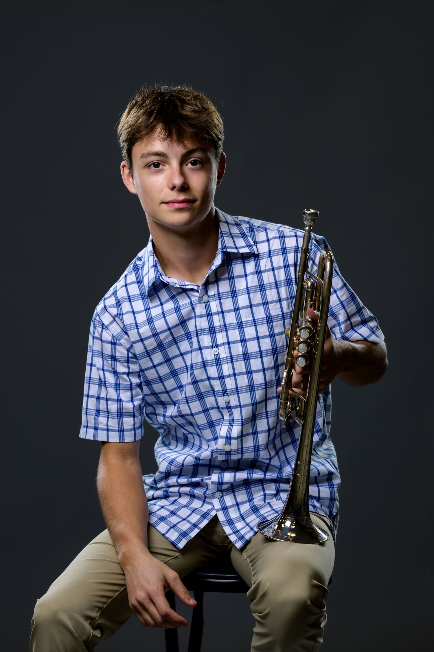 Student musician poses with clarinet in blue checkered shirt against dark studio background.