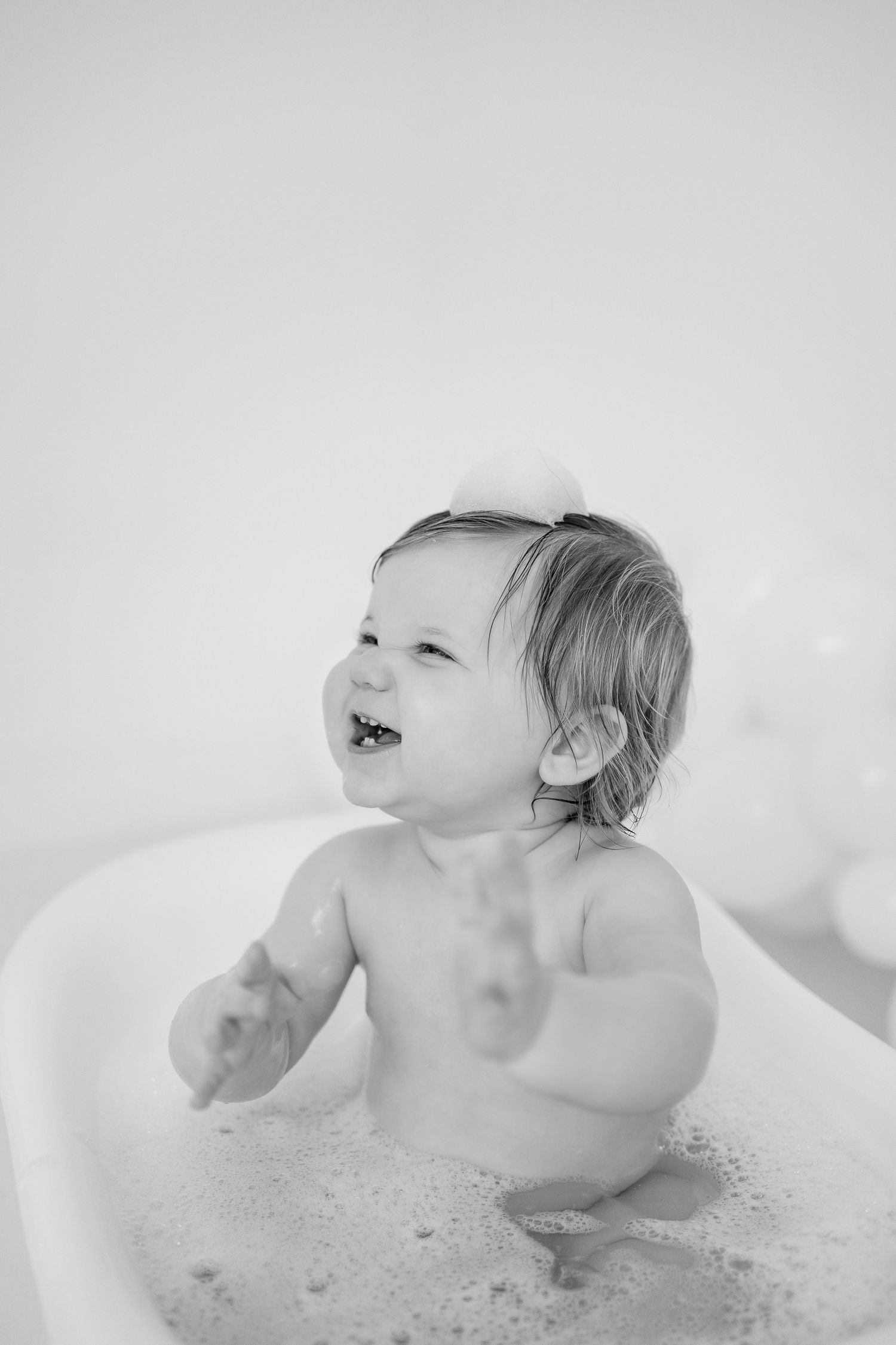 A baby plays and splashes in a bubble bath while looking up with joy in a black and white photo series.