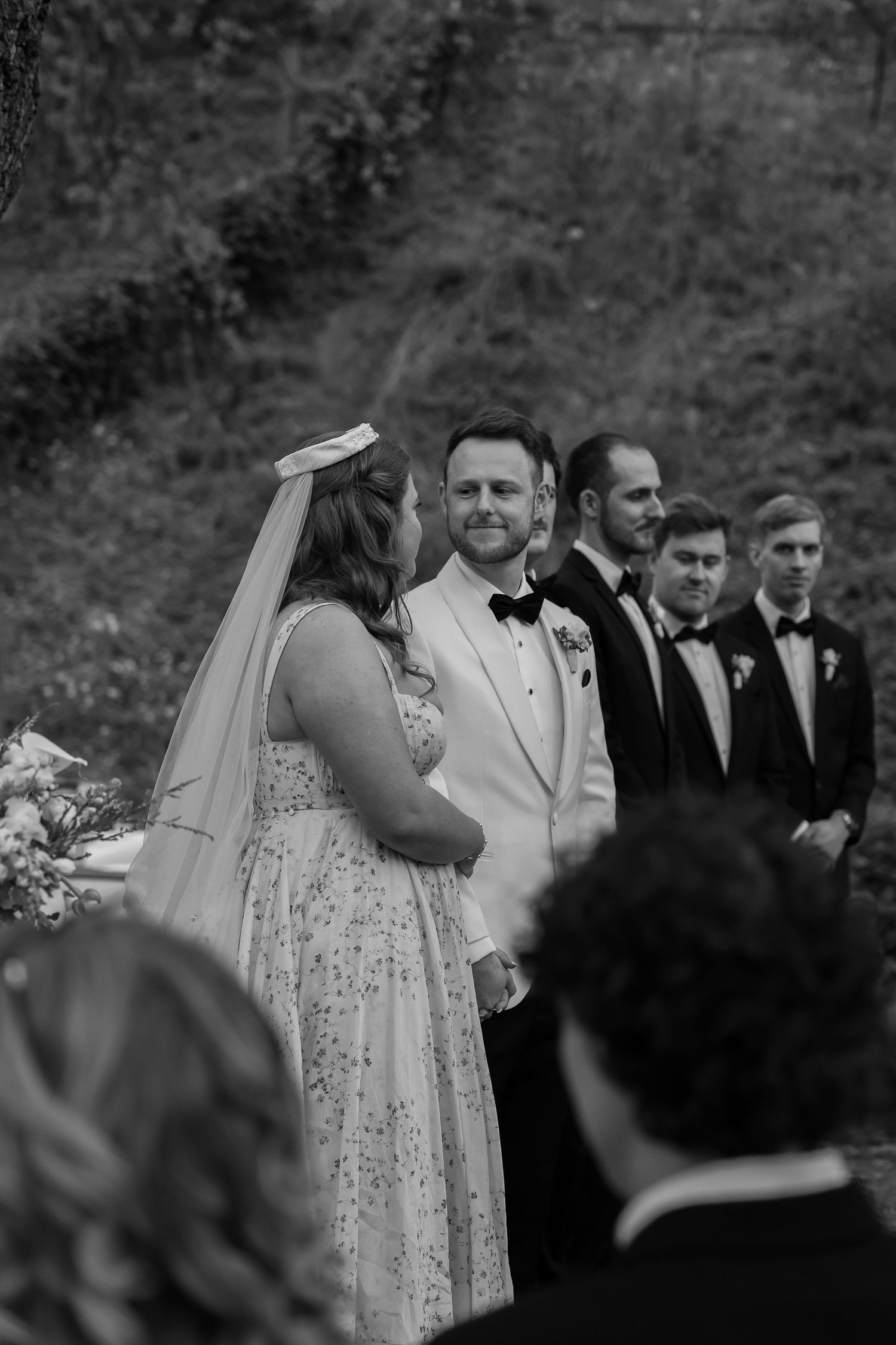 Bride and groom stand together during their outdoor ceremony, surrounded by their wedding party and lush greenery. in black and white