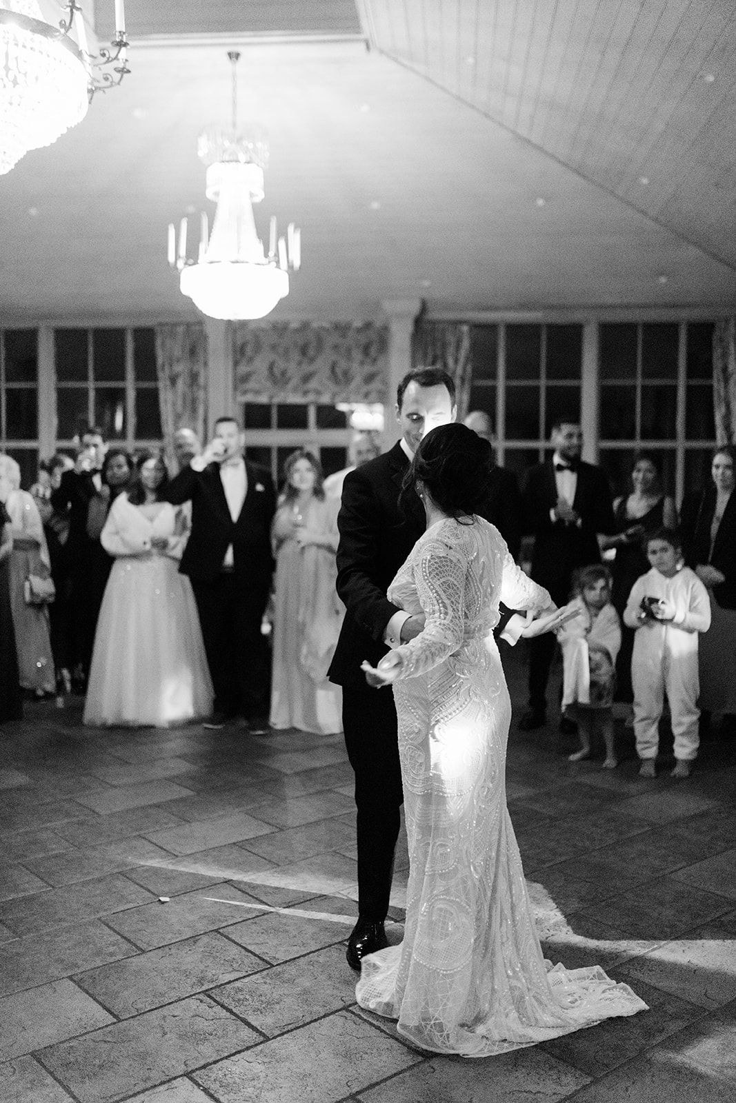 A couple shares their first dance at an elegant wedding reception under crystal chandeliers while guests look on.