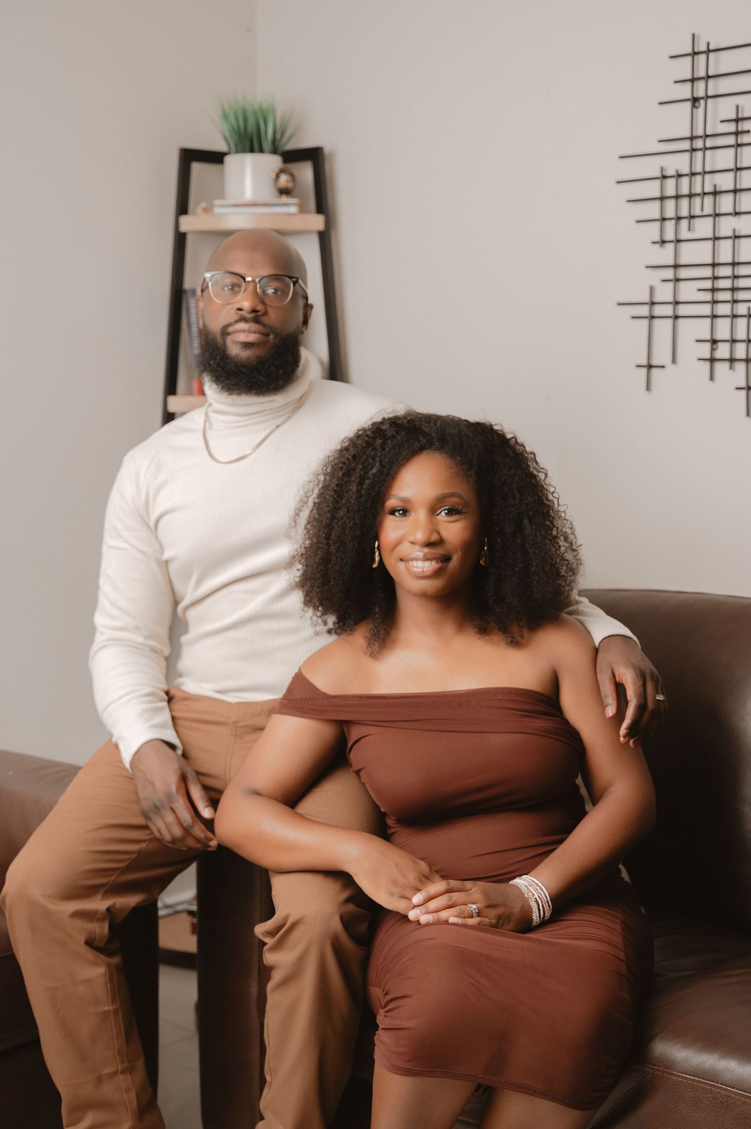 A couple poses together in brown coordinated outfits against a neutral wall for a romantic photoshoot series.